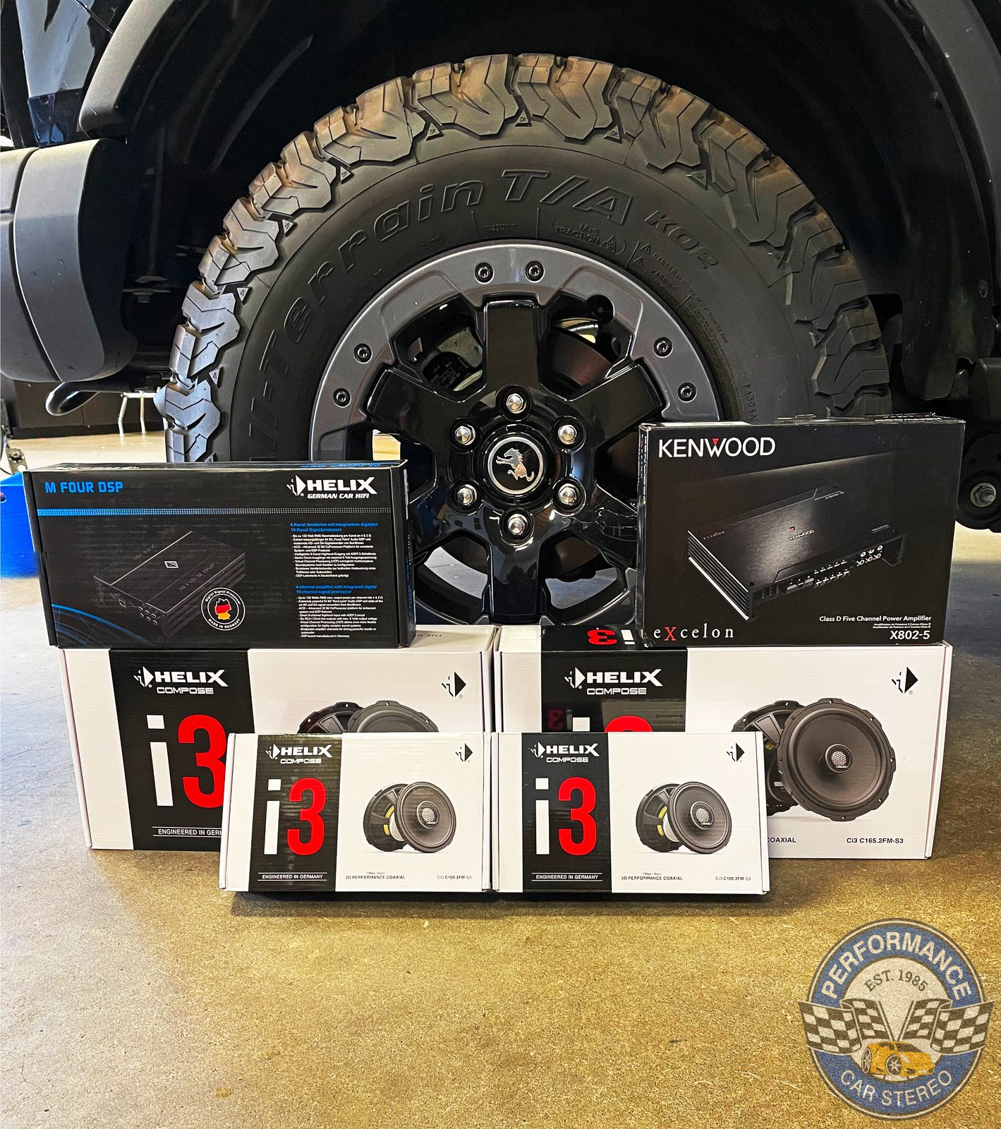 Helix and Kenwood boxes laid out on the shop floor beside the Bronco's wheel: an M FOUR DSP, a Kenwood X802-5 amplifier and four boxes of Helix Compose i3 speakers