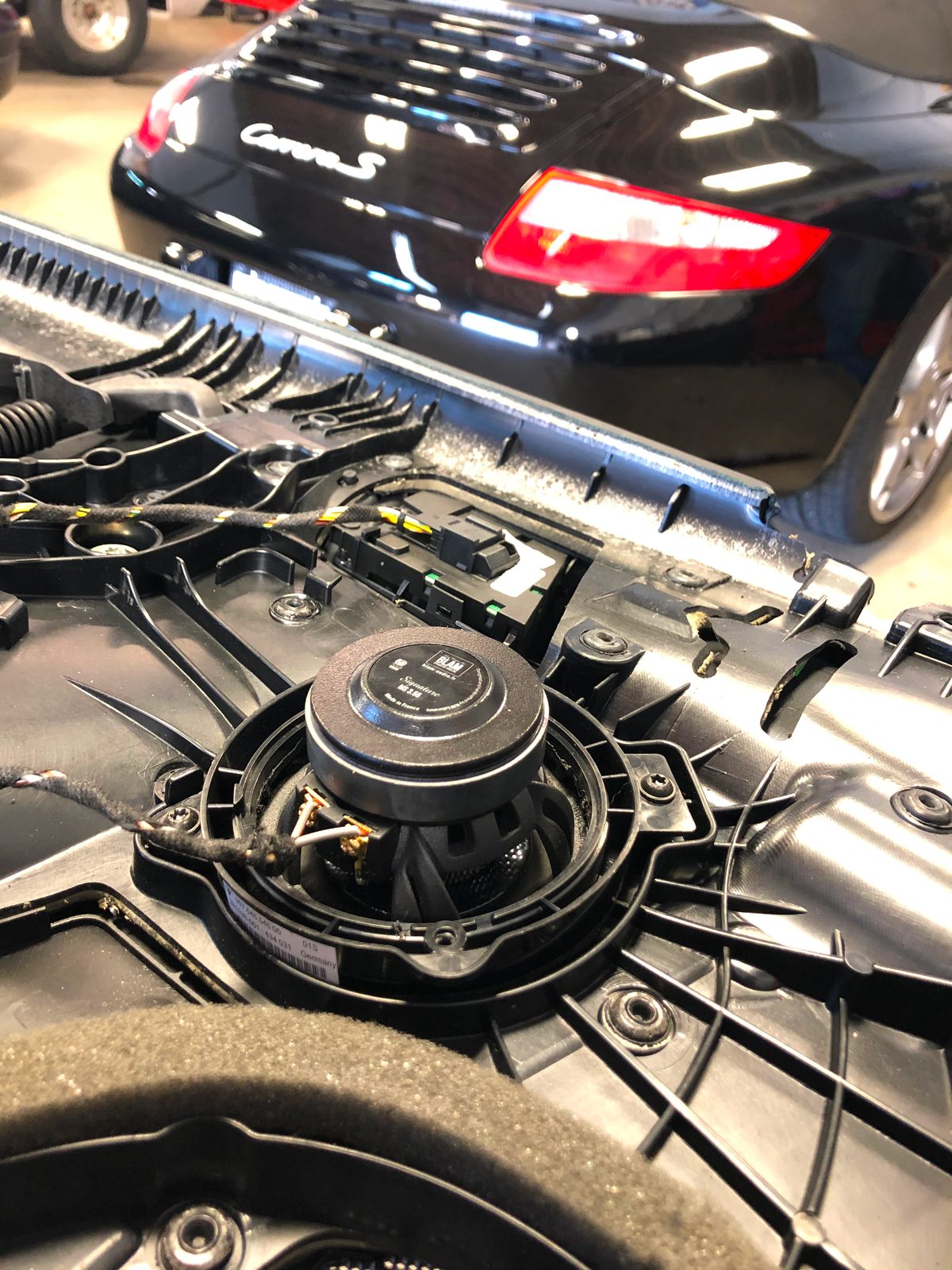 A BLAM Signature driver fitted into a factory Porsche speaker frame on a removed panel, with the black Carrera S in the background