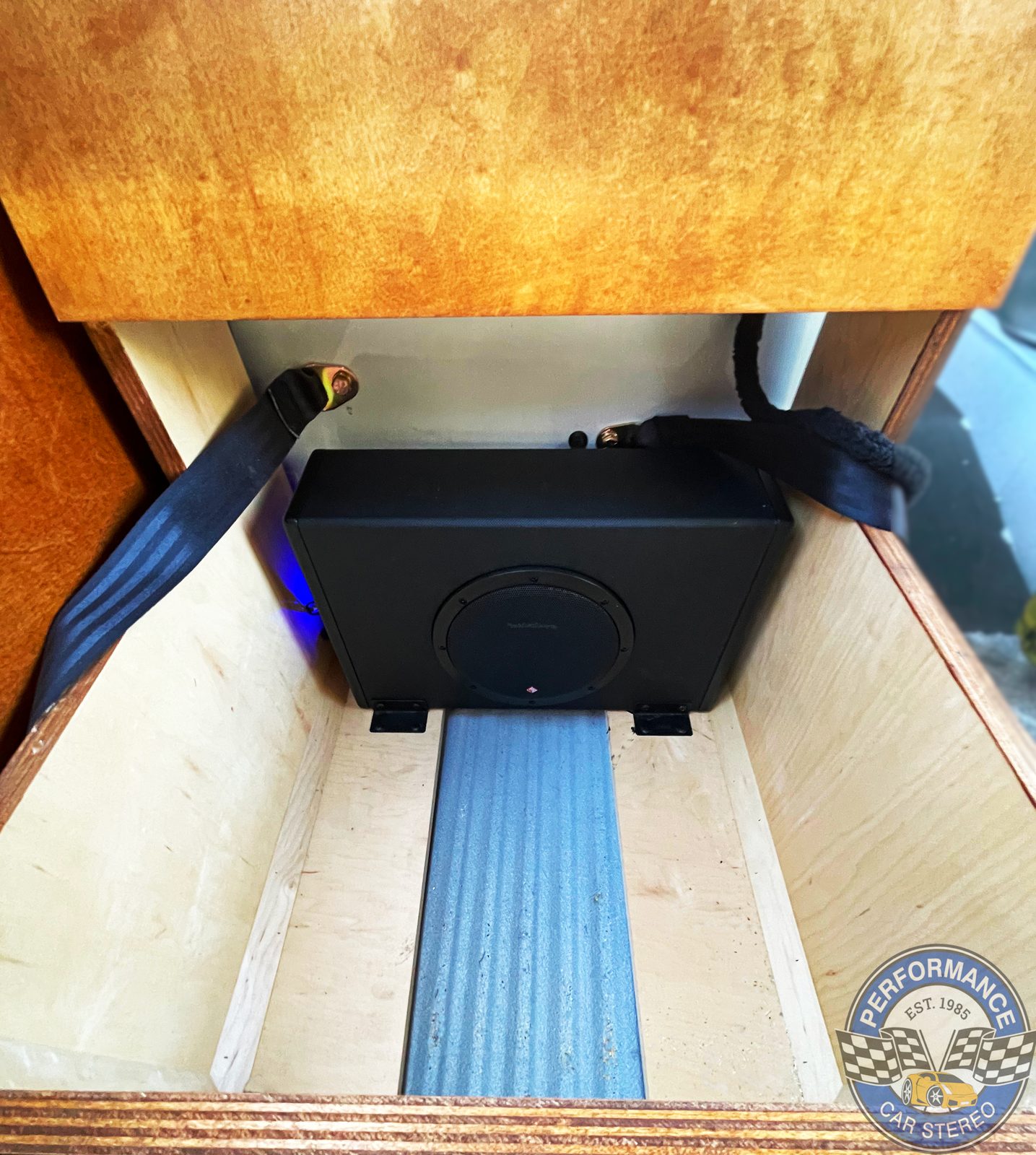 A Rockford Fosgate powered subwoofer enclosure strapped inside the plywood box beneath the front bench seat, with the hinged wooden lid propped open
